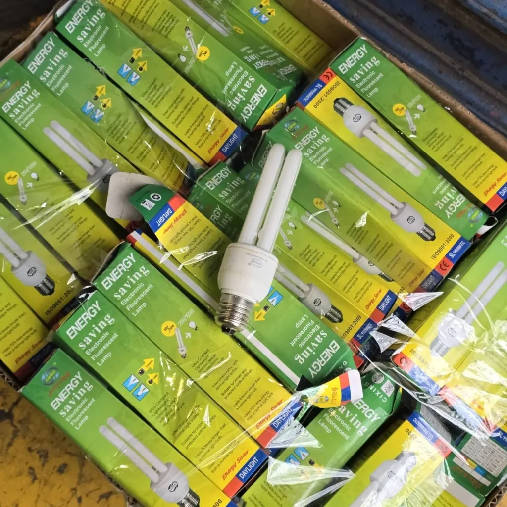 Caja llena de lámparas fluorescentes electrónicas de bajo consumo en empaques verdes y amarillos, con una bombilla suelta visible. by Remates Narvaja