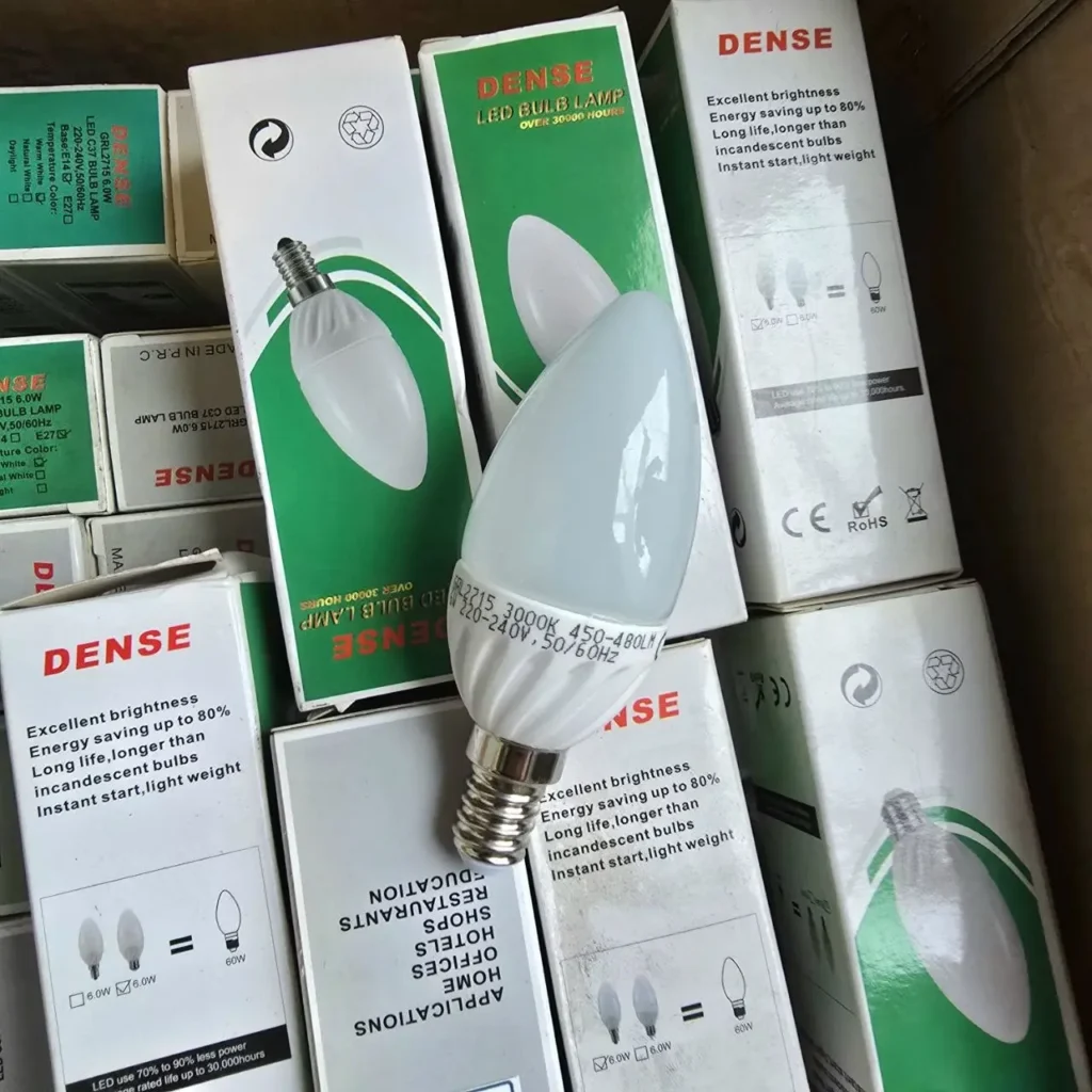 Bombilla LED tipo vela DENSE blanca con base E14, rodeada de cajas apiladas de bombillas LED DENSE, destacando ahorro de energía (6.0W equivalente a 60W) y larga vida útil. by Remates Narvaja