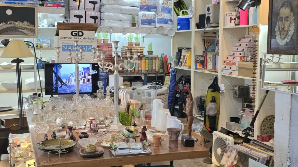 Tienda o almacén de remates con mesa llena de cristalería, vajilla y artículos para el hogar, rodeada de estanterías con by Remates Narvaja