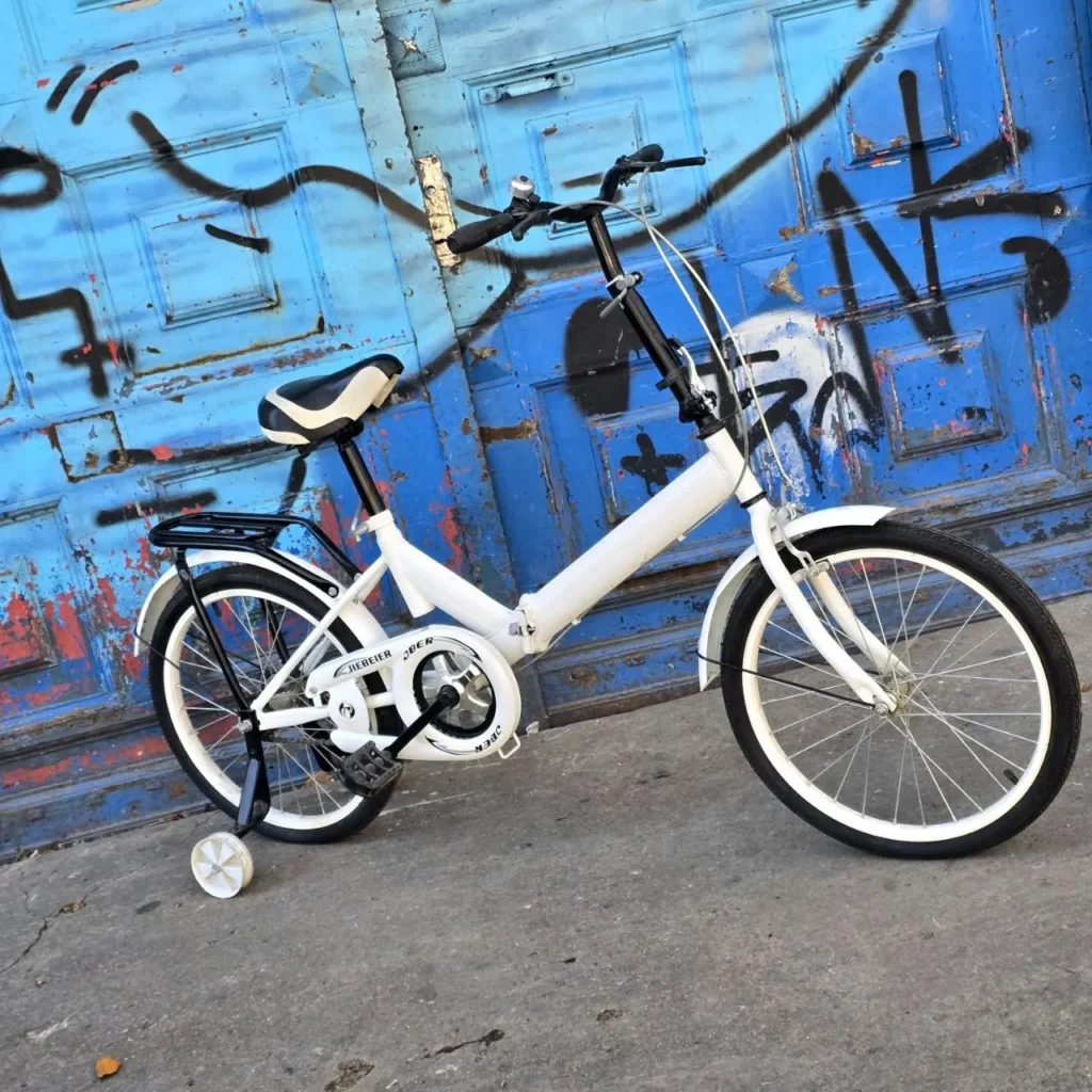 Bicicleta blanca para niños con rueditas de entrenamiento frente a una puerta azul grafiteada. by Remates Narvaja