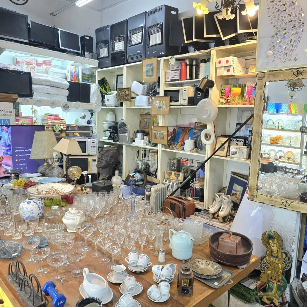 Tienda de segunda mano con estanterías llenas de electrónica, electrodomésticos, libros y decoración, y una mesa con cristalería by Remates Narvaja