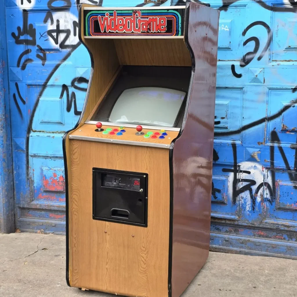 Máquina arcade retro de madera con letrero "videogame" y controles, frente a una pared azul con grafitis. by Remates Narvaja