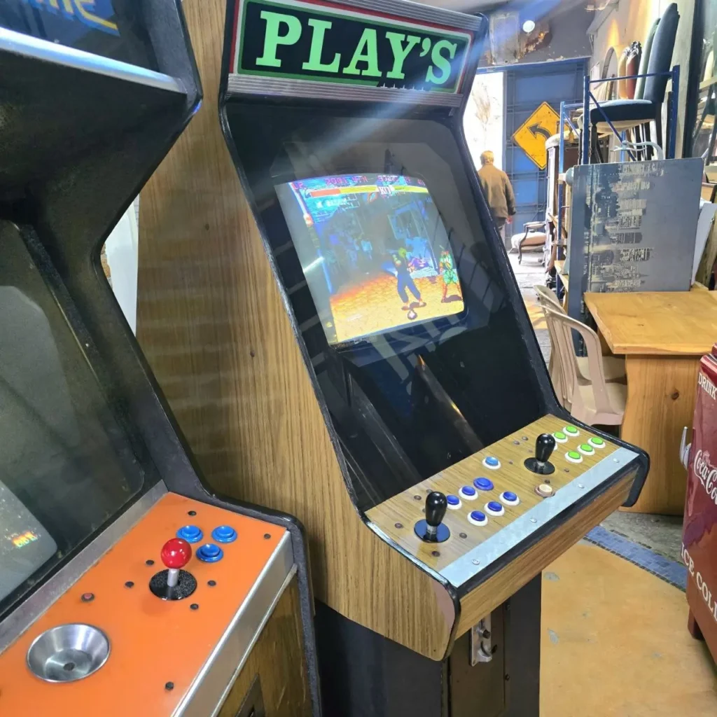 Dos máquinas arcade vintage con joysticks y botones, una mostrando un juego de lucha, en una tienda. by Remates Narvaja