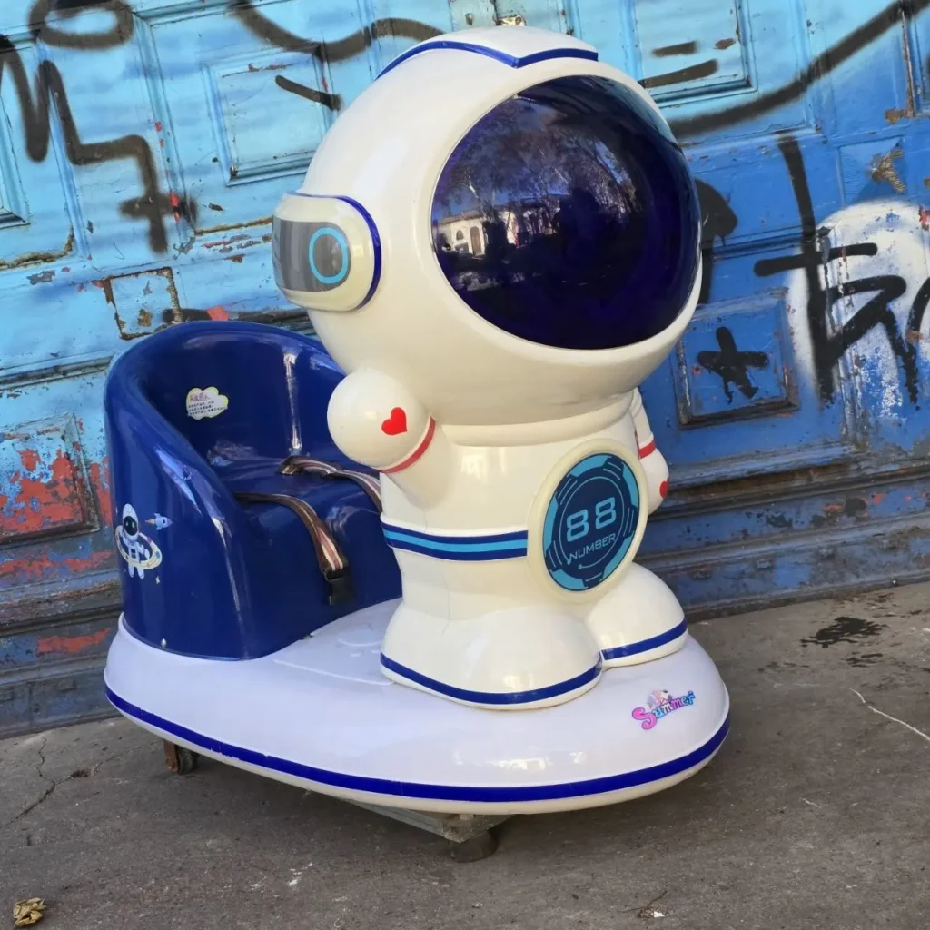 Máquina recreativa infantil de astronauta blanca y azul con asiento, frente a una pared azul con grafitis. by Remates Narvaja