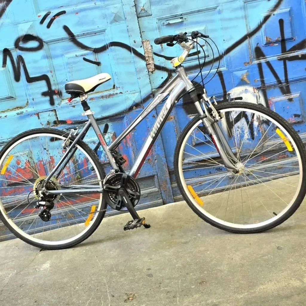 Bicicleta híbrida gris Winner con suspensión delantera y sillín blanco, frente a una pared azul con grafitis. by Remates Narvaja