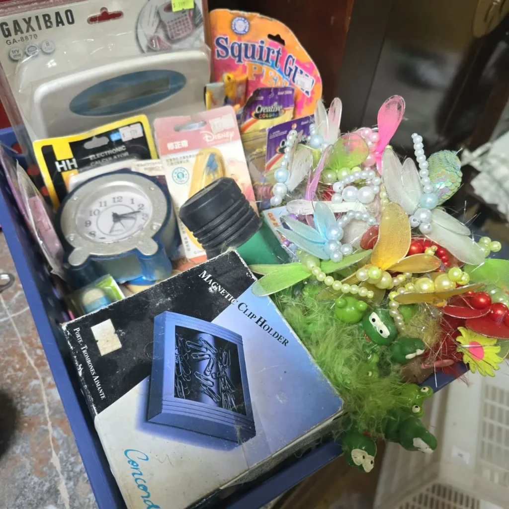 Caja azul llena de artículos variados como un reloj despertador, calculadora, sujetapapeles magnético, adornos de libélulas y ranas by Remates Narvaja