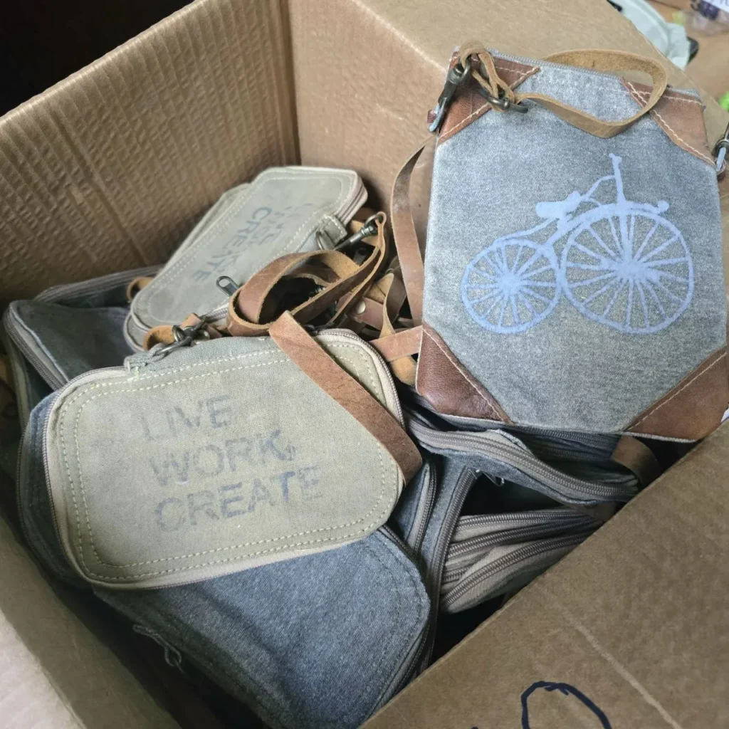 Caja llena de bolsas de lona gris con detalles de cuero, una con diseño de bicicleta y otra con texto "LIVE WORK CREATE". by Remates Narvaja