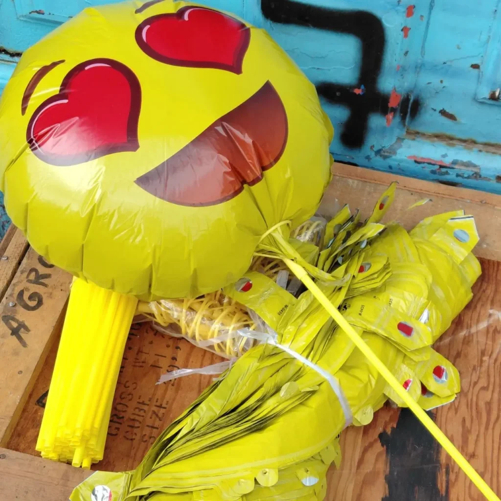 Globo emoji amarillo de ojos de corazón inflado, junto a una pila de globos desinflados y palos amarillos para fiesta. by Remates Narvaja
