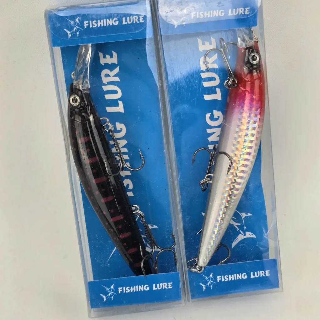 Dos señuelos de pesca en su empaque, uno negro con rayas rojas y otro plateado holográfico con cabeza roja. by Remates Narvaja