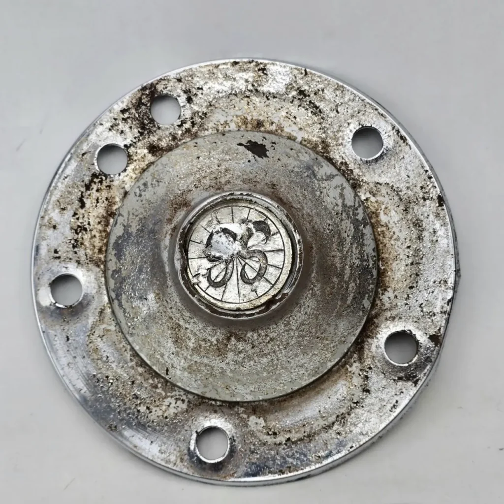 Tapacubo metálico circular corroído con emblema "GS" y agujeros de fijación. by Remates Narvaja