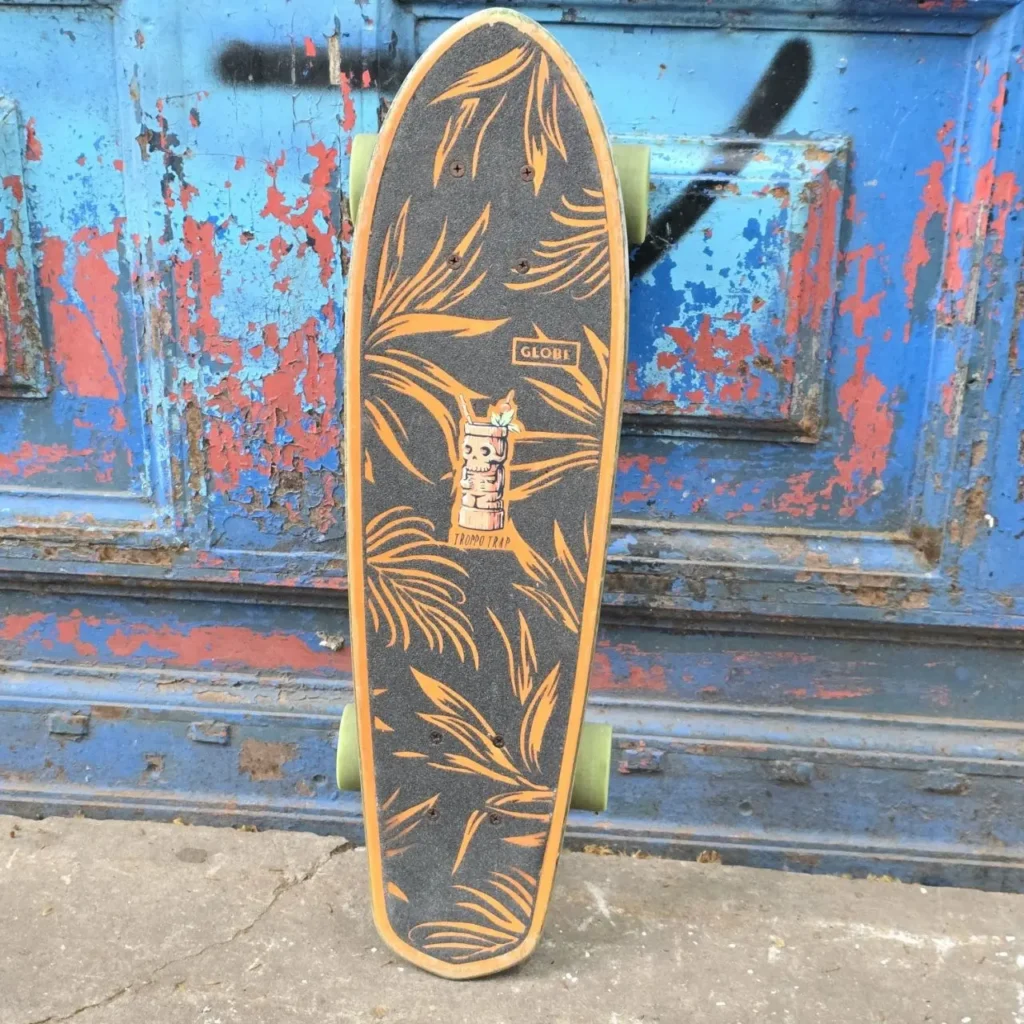 Longboard Globe con diseño tropical de hojas y tiki, negro y naranja, apoyado en una puerta azul desgastada. by Remates Narvaja